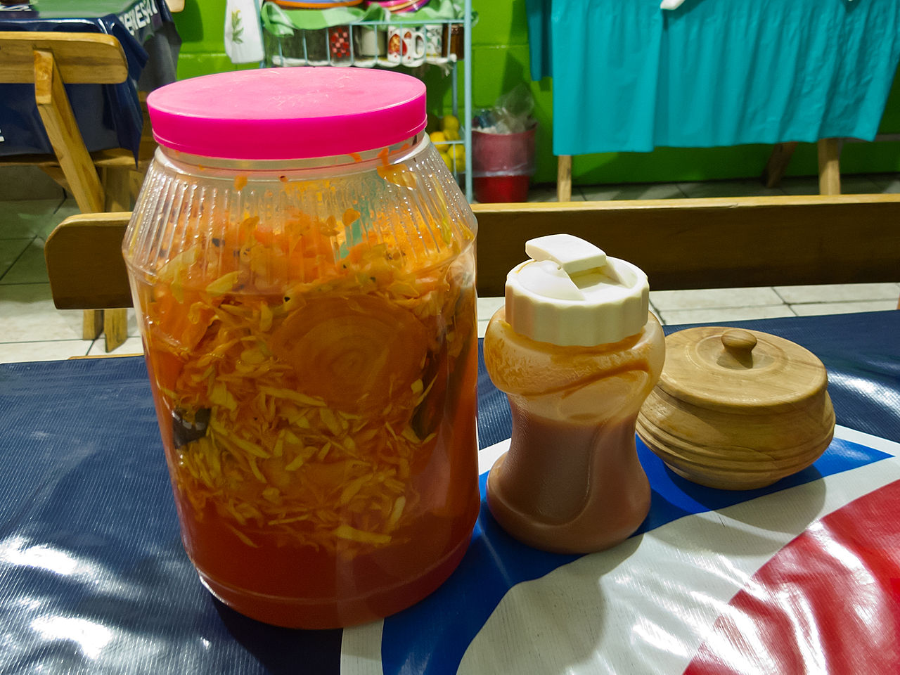 Condiments for Pupusas, typical for El Salvador (This photo here was taken in Olocuilta, La Paz, El Salvador). Large container at left is the vingared cabbage (curtido), tomato sauce in the middle. (Wooden sugar bowl at right has sugar for coffee)