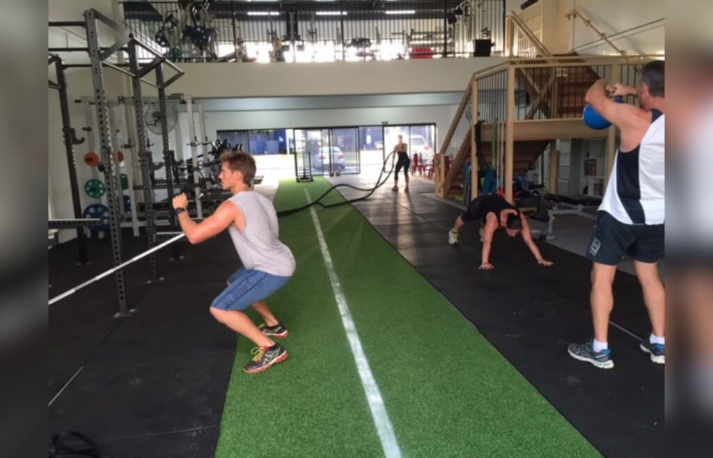 A gym where various forms of strength training are being practiced. From left: overhead presses, battle ropes, planking, and kettlebell raises.