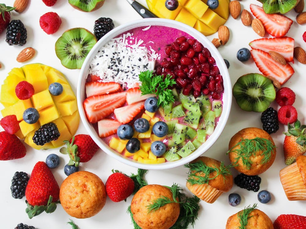Smoothie bowl with kiwi, strawberries, pomegranate, mango, blueberries, and coconut surrounded by fresh fruit and muffins for smart snacking ideas.